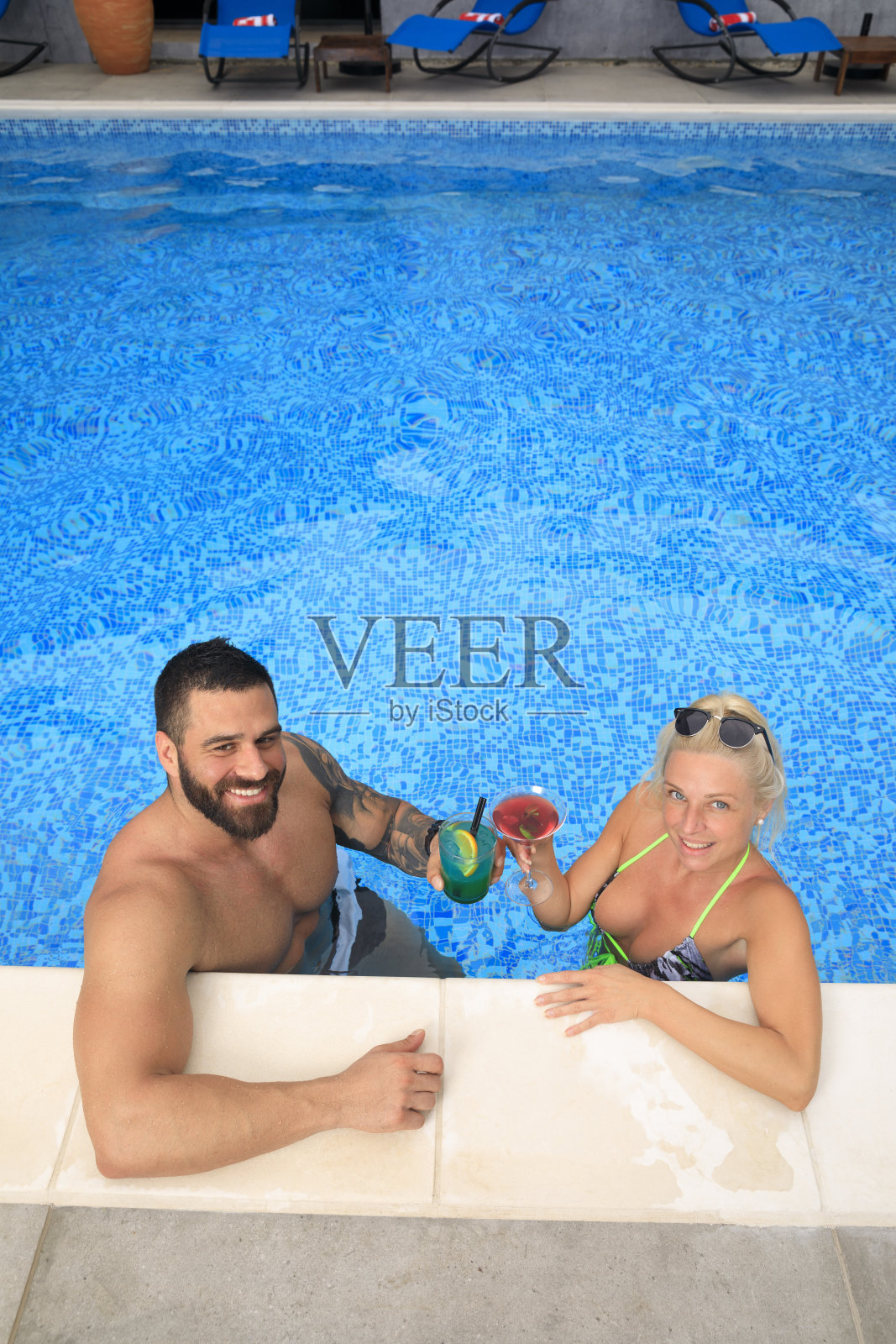 一对相爱的情侣在泳池边的夏季派对，在游泳池的边缘，年轻的女人和男人喝鸡尾酒，享受夏天的乐趣。暑假的概念照片摄影图片
