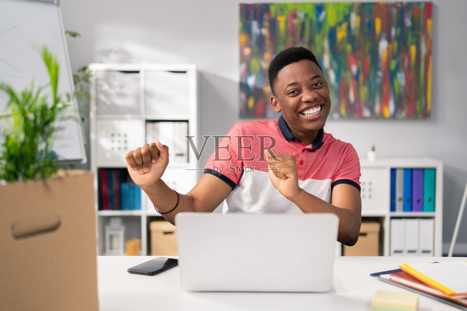 身穿t恤、皮肤黝黑的男子满意于售出的服务产品，满意于每月的利润，坐在电脑前胜利地跳舞，紧握拳头望着相机，微笑着照片摄影图片