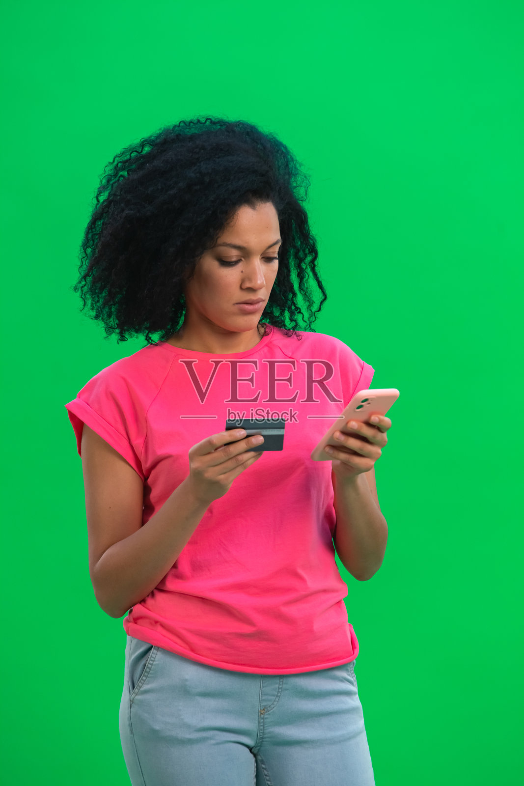 年轻的非洲裔美国女性肖像用信用卡和手机在网上购物。一名身穿粉色t恤、一头卷发的黑人女子在演播室的绿色屏幕上摆姿势。近距离照片摄影图片