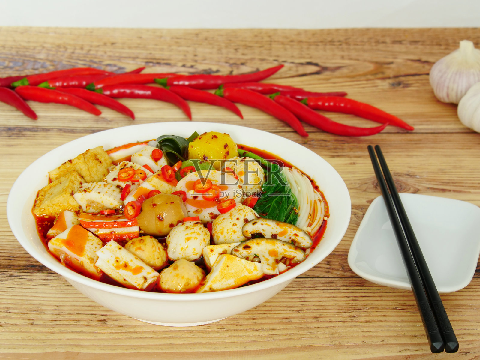 木桌上特写一碗美味火锅配料:香菇、肉丸、海带、金菇、豆腐泡等。碗旁边有红辣椒、大蒜和一双筷子。照片摄影图片