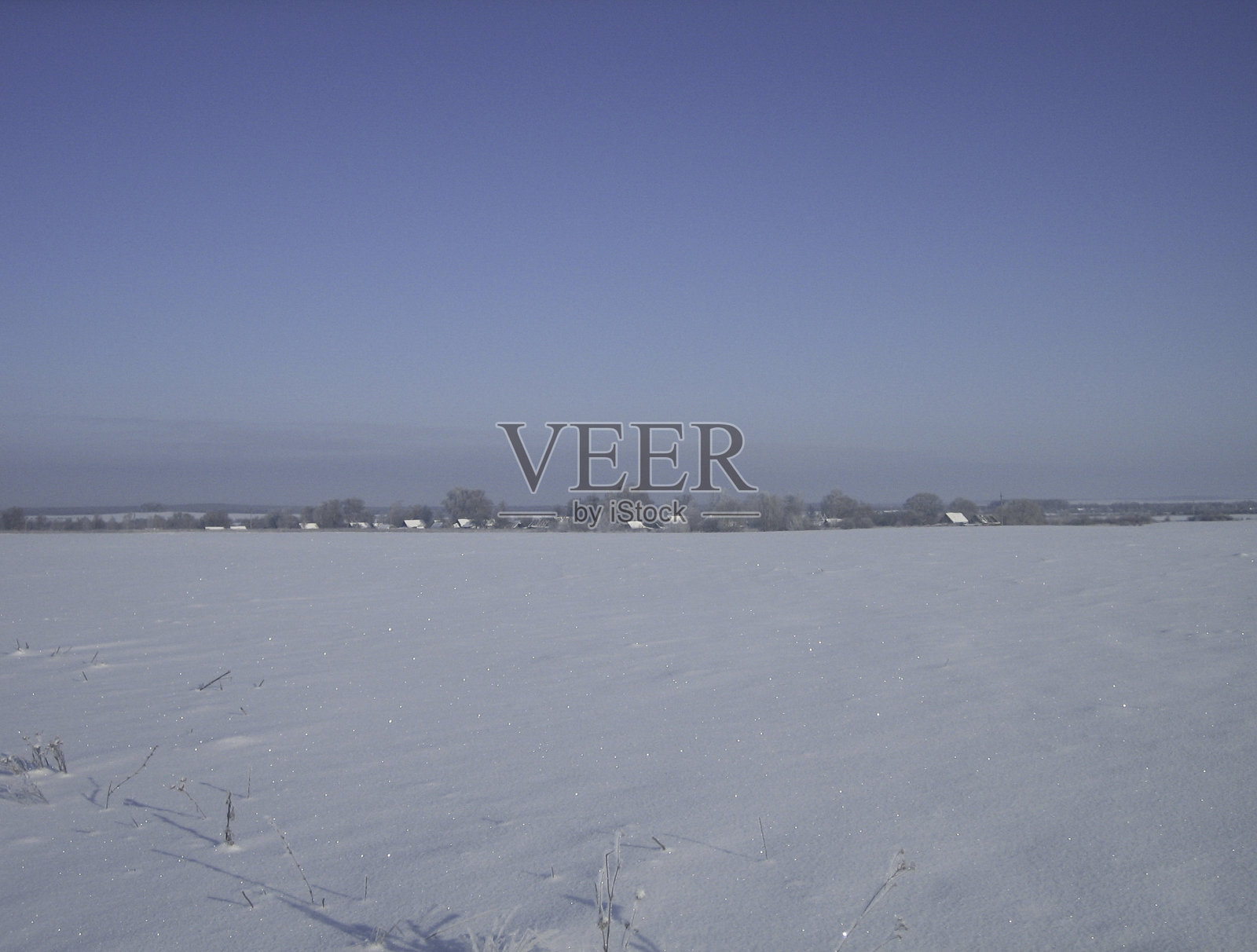 在一个晴朗的霜天被雪覆盖的乡村平地。在远处，你可以看到村庄的房子。照片摄影图片