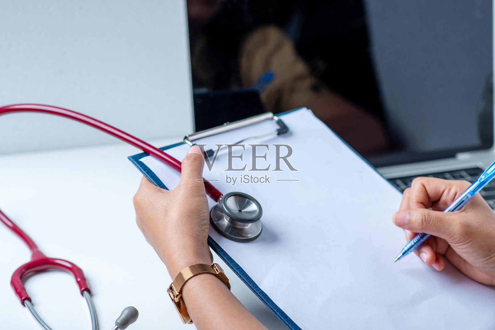 医生用钢笔写下疾病的症状。白色的桌子上有一个听诊器和一台电脑。照片摄影图片