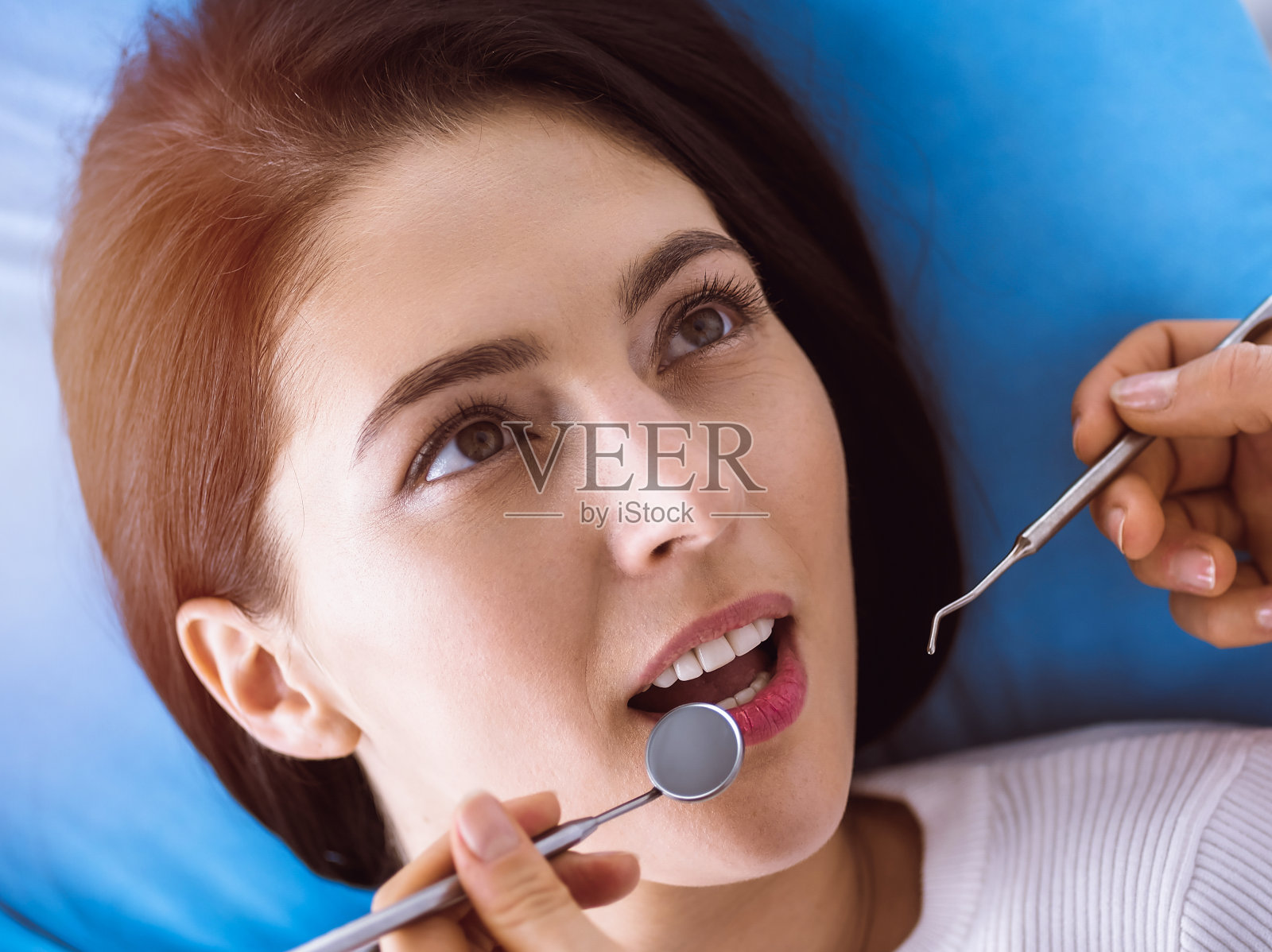 微笑的黑发女人正在检查牙医在阳光牙科诊所。医生的手拿着牙科器械靠近病人的嘴。健康牙齿和医学概念照片摄影图片