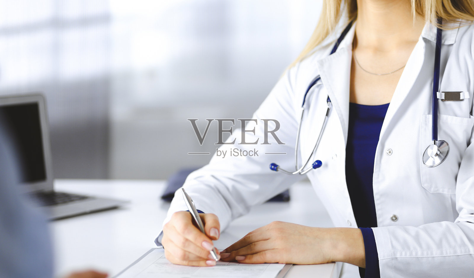 一位不知名的女医生坐在诊所橱柜的桌子旁，在她的病历上写下病人的感冒症状。女医生正在用写字板和听诊器照片摄影图片