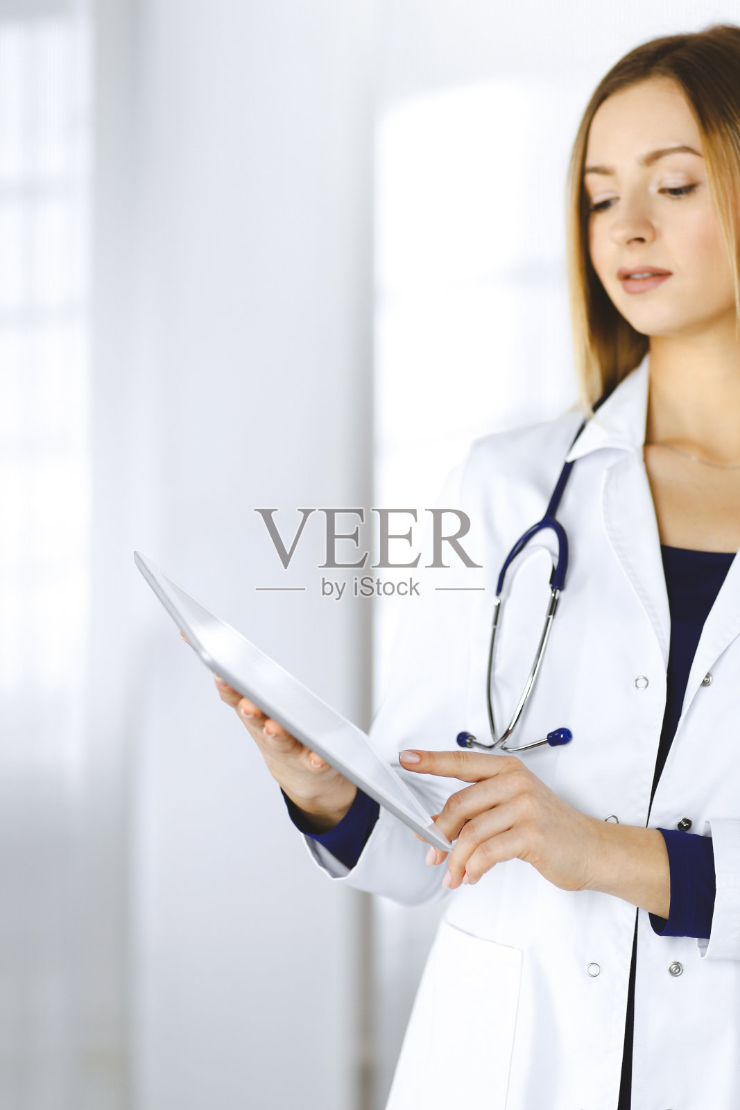 一位年轻的女医生站在诊所里，手里拿着一台平板电脑。友好的女医生的肖像与听诊器在工作。医院完善的医疗服务医学的概念照片摄影图片