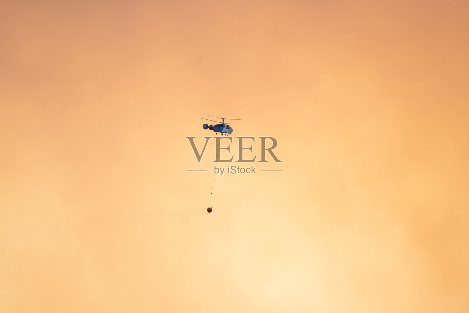 空中消防在黄色多云的天空背景中带着水篮的飞行直升机。照片摄影图片