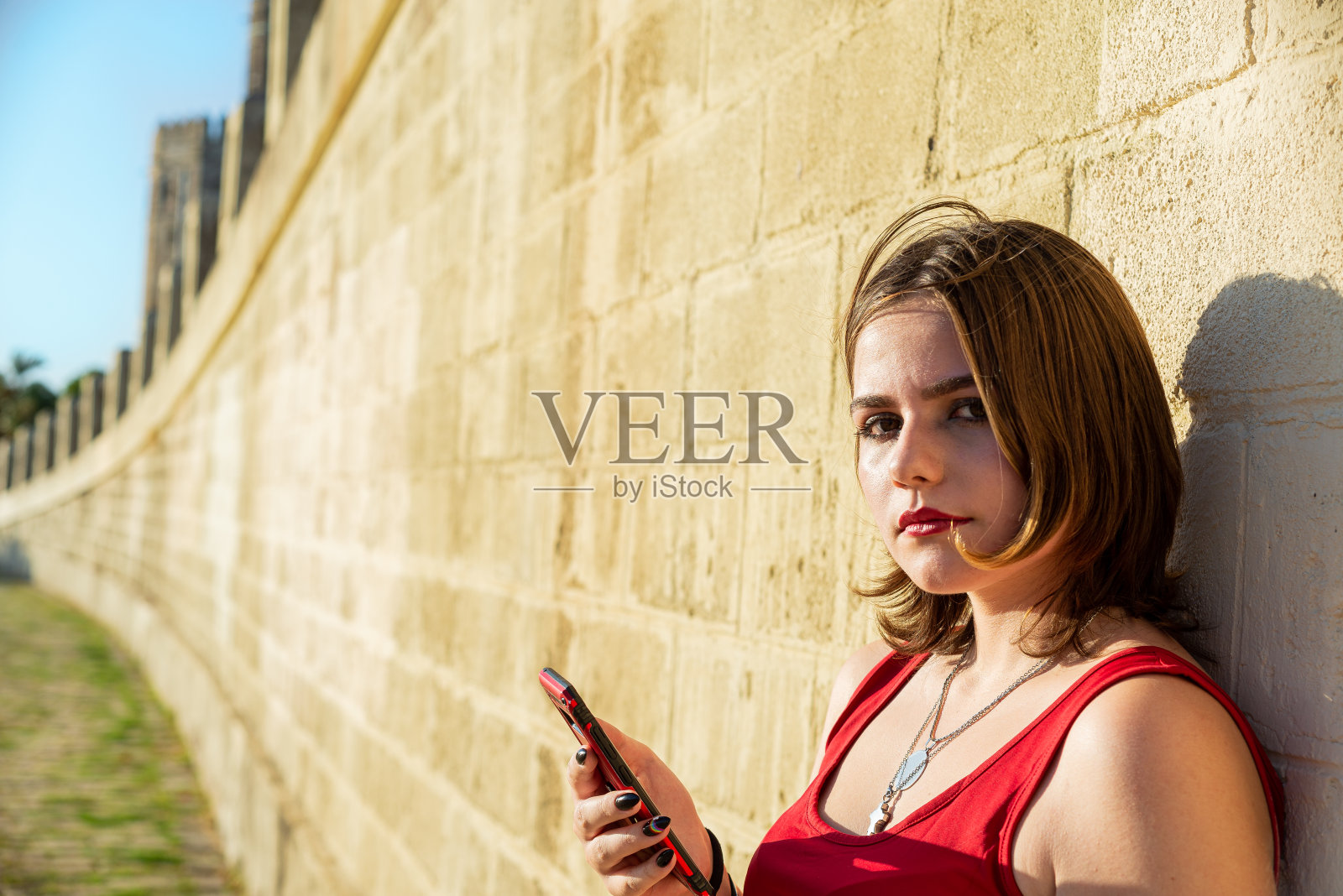 年轻的短发女子拿着智能手机看着镜头，背景是一堵黄色的墙，墙上有阳光照片摄影图片