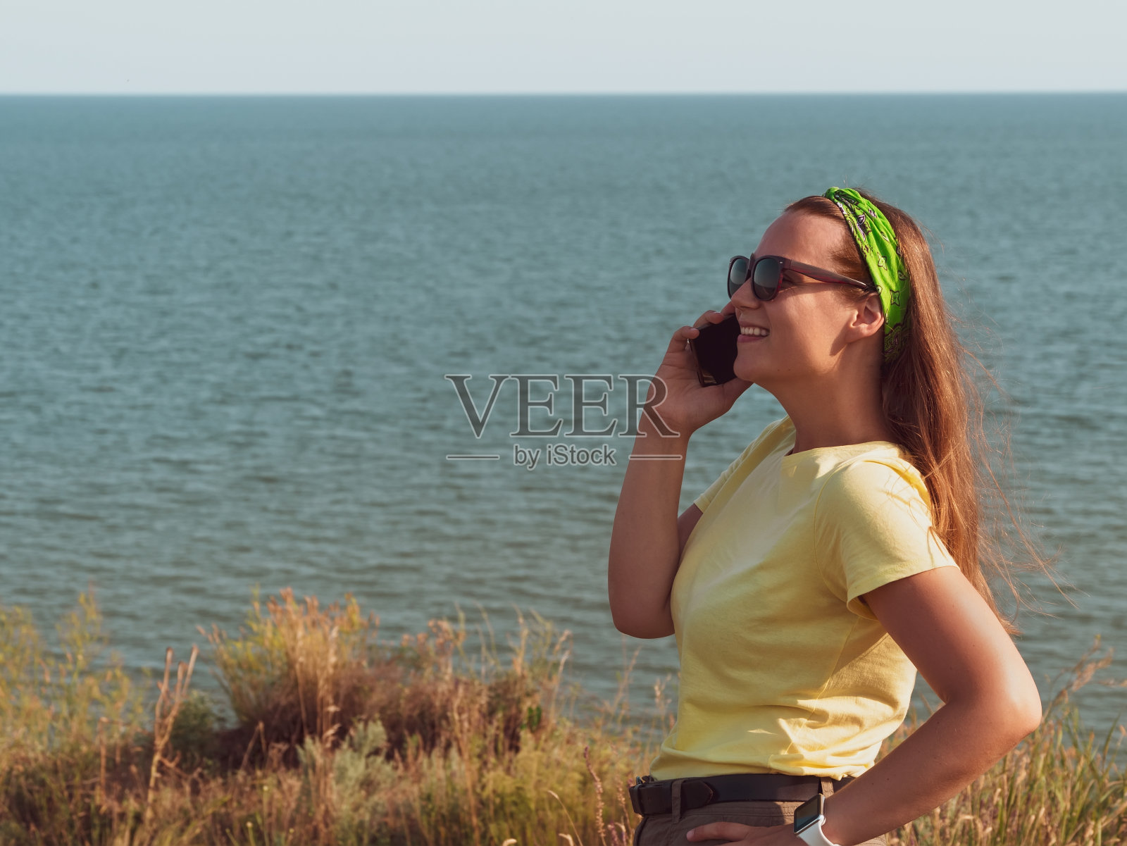 年轻自信的女人穿着黄色t恤，手里拿着智能手机，在海上自拍。时尚的千禧一代地道女孩照片摄影图片