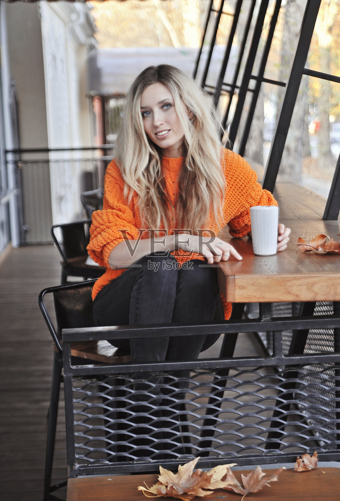 美丽的年轻女子在橙色毛衣摆姿势咖啡因杯拿铁和秋天的叶子。照片摄影图片
