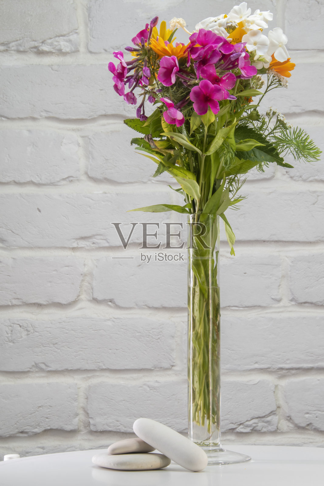 在白色砖墙的背景下，白色桌子上放着一束放在玻璃花瓶里的鲜花。照片摄影图片