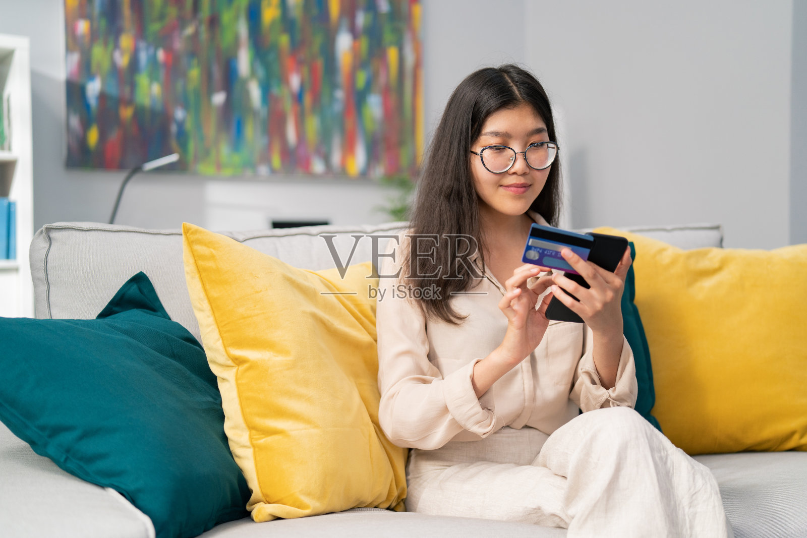 专注的亚洲美女美女在客厅的沙发上，她手里拿着电话和银行、信用卡，女人正在网上购物、付款，输入号码安全码照片摄影图片