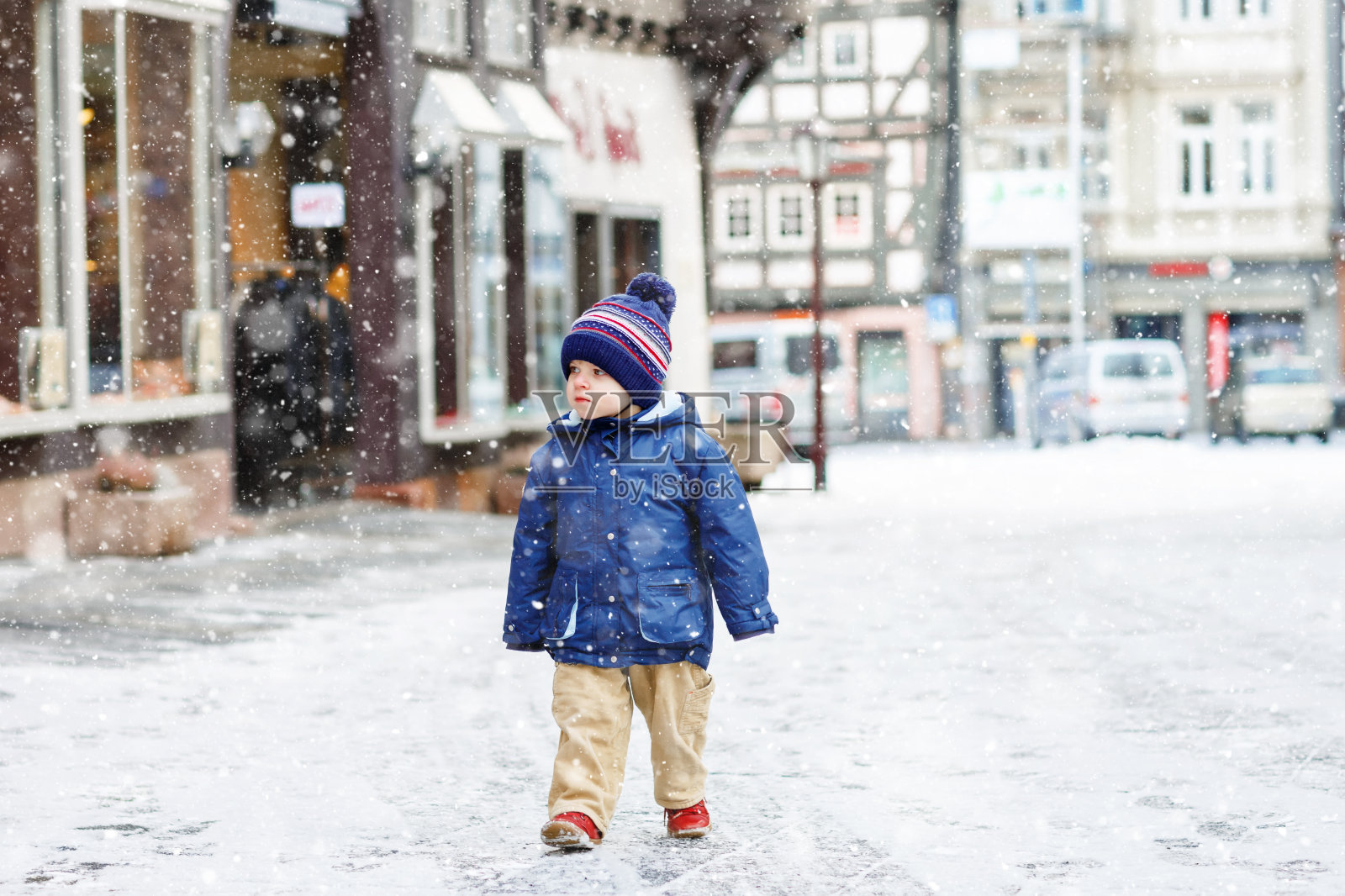 一个蹒跚学步的小男孩在下雪的时候走过被雪覆盖的城市。可爱快乐的孩子穿着冬衣在户外玩耍，在寒冷的日子里有雪在街上。照片摄影图片