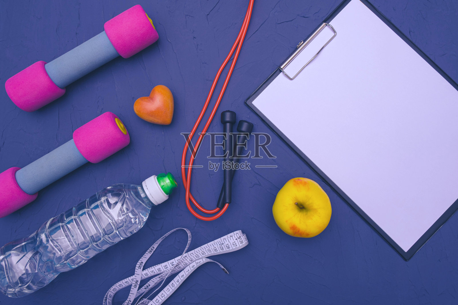 运动器材——哑铃，一个苹果，一瓶水和一根跳绳，一张记录成绩的白纸，蓝色背景照片摄影图片