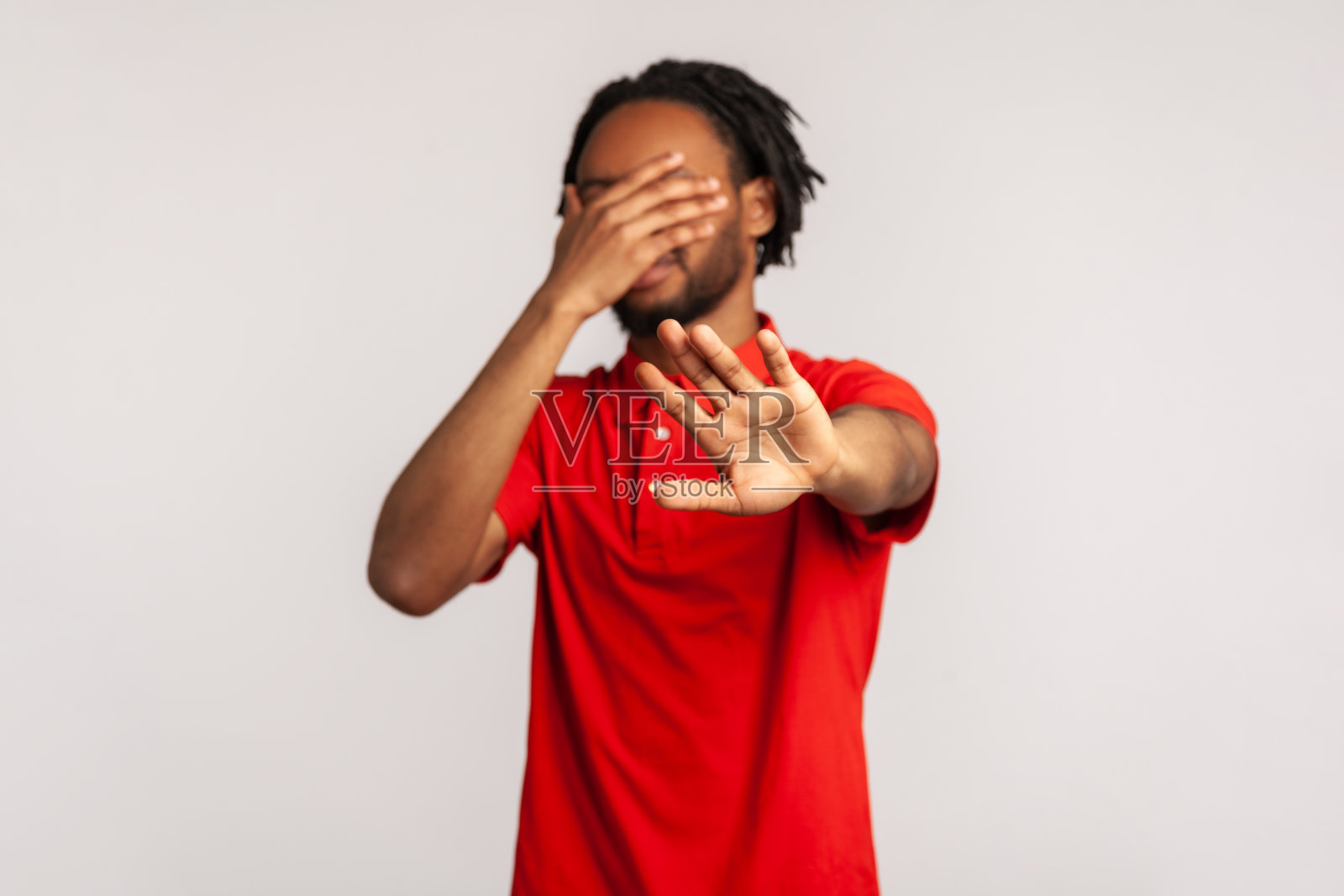 停止，我不想看这个。留着脏辫的年轻络腮胡男子穿着红色休闲T恤，遮住脸，做出停止手势。照片摄影图片