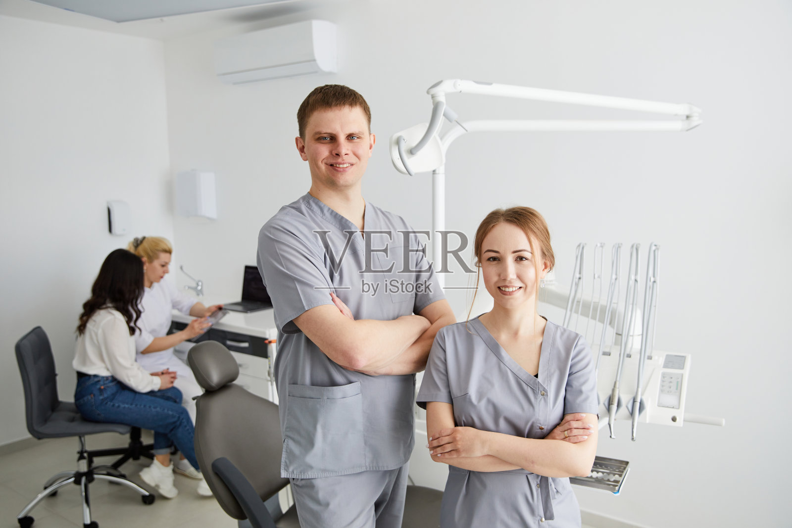 两名牙医——一男一女站在办公室里，后面坐着一位病人和一名助手。牙医的办公室照片摄影图片