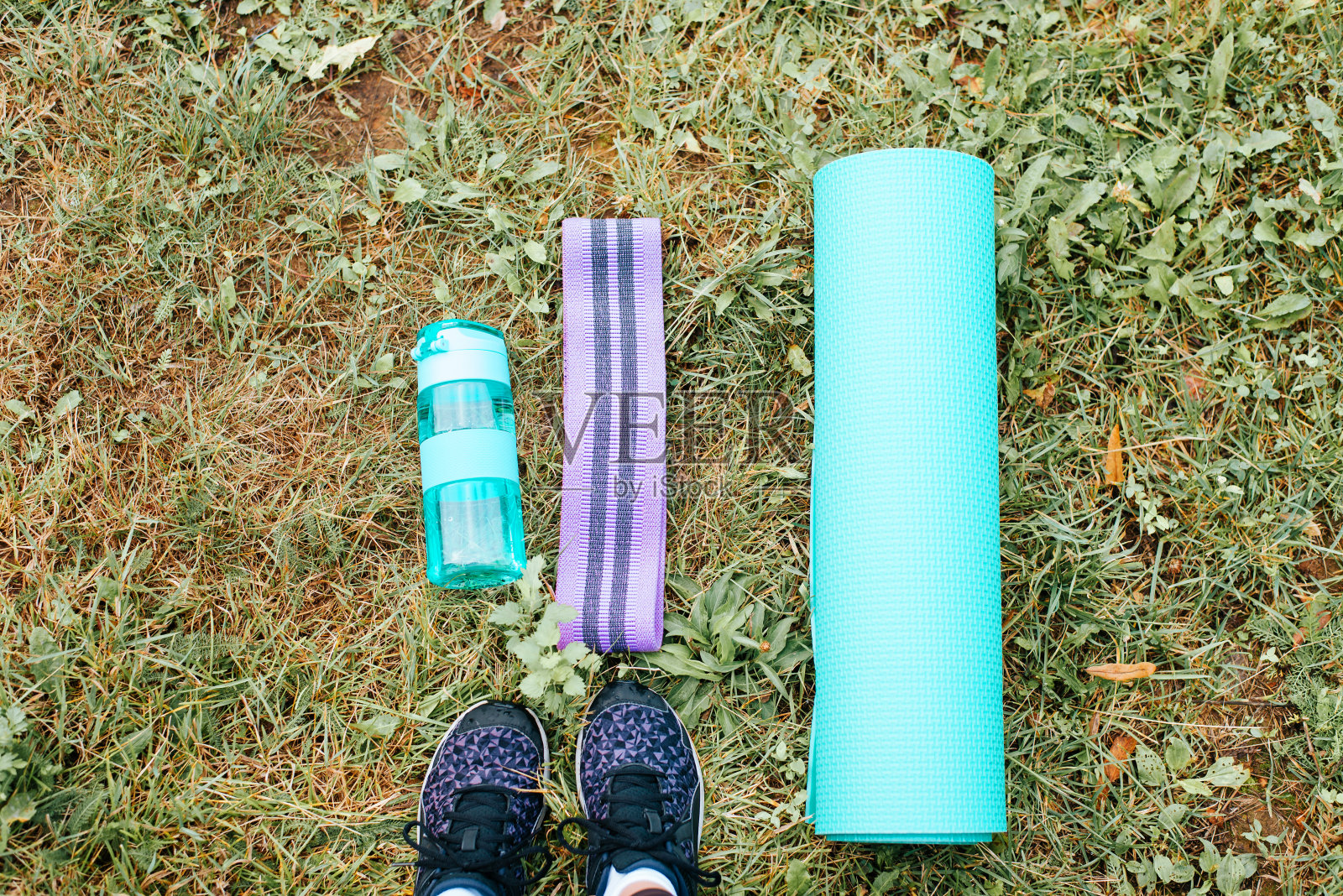 健康积极的生活方式，运动理念。瑜伽垫，弹性膨胀器，水瓶和女性腿在绿色草地上的运动鞋顶部视图，户外。户外训练器材的第一人称视角照片摄影图片