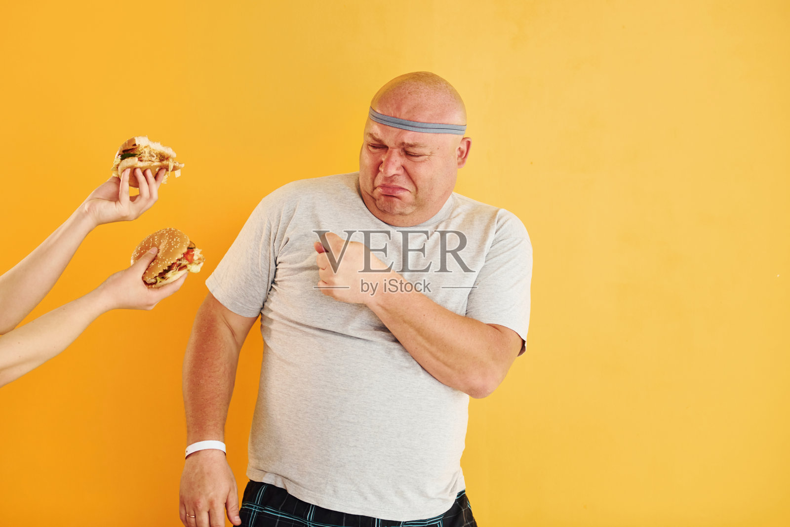手拿汉堡诱惑男人。有趣的超重男子在运动头领带是在黄色背景照片摄影图片
