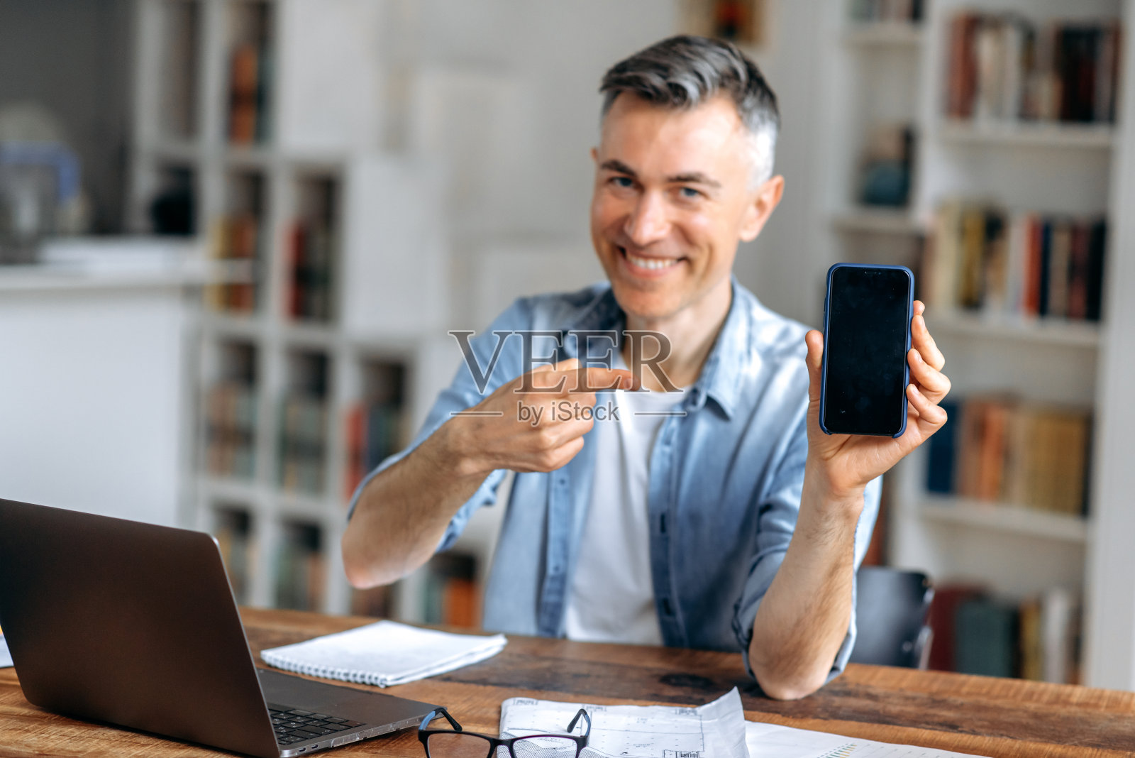 成功的自信有吸引力的成熟白种人男子，经理，经纪人或自由职业者，在笔记本电脑后面远程工作，手指在智能手机，看着相机，微笑照片摄影图片