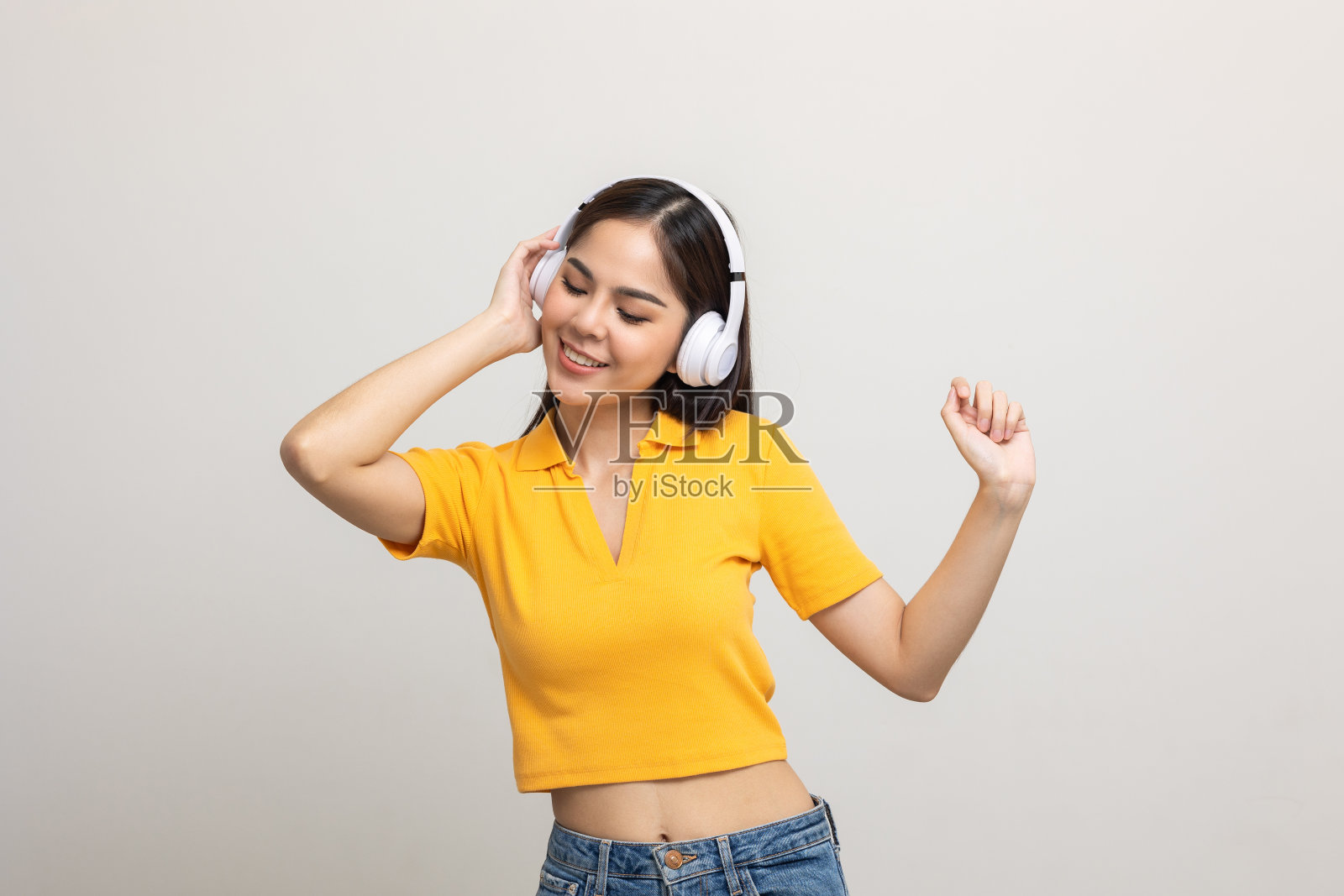 恐怖的亚洲少女听音乐与白色耳机跳舞在孤立。漂亮的年轻女子在黄色衬衫手触摸无线耳机与音乐在房间里玩。照片摄影图片