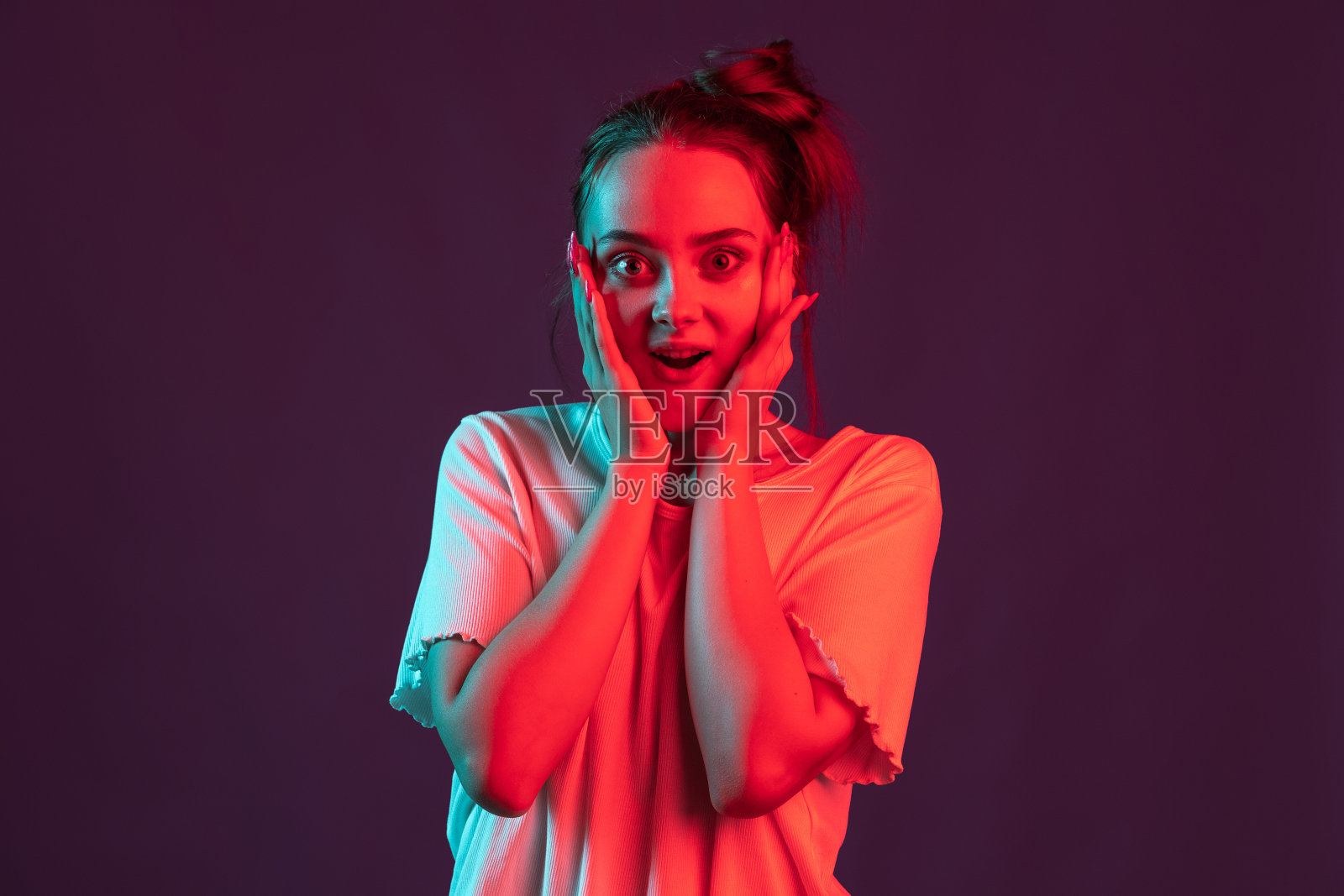 美丽女孩的肖像在休闲服装孤立在暗紫色的工作室背景在红色霓虹灯，滤镜。人类情感的概念，面部表情。照片摄影图片