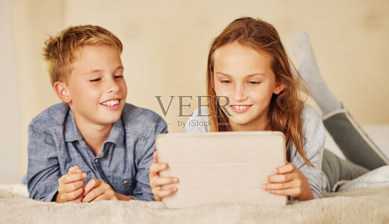 两个小孩一起躺在家里的床上，一边玩电子平板电脑，一边亲密无间照片摄影图片