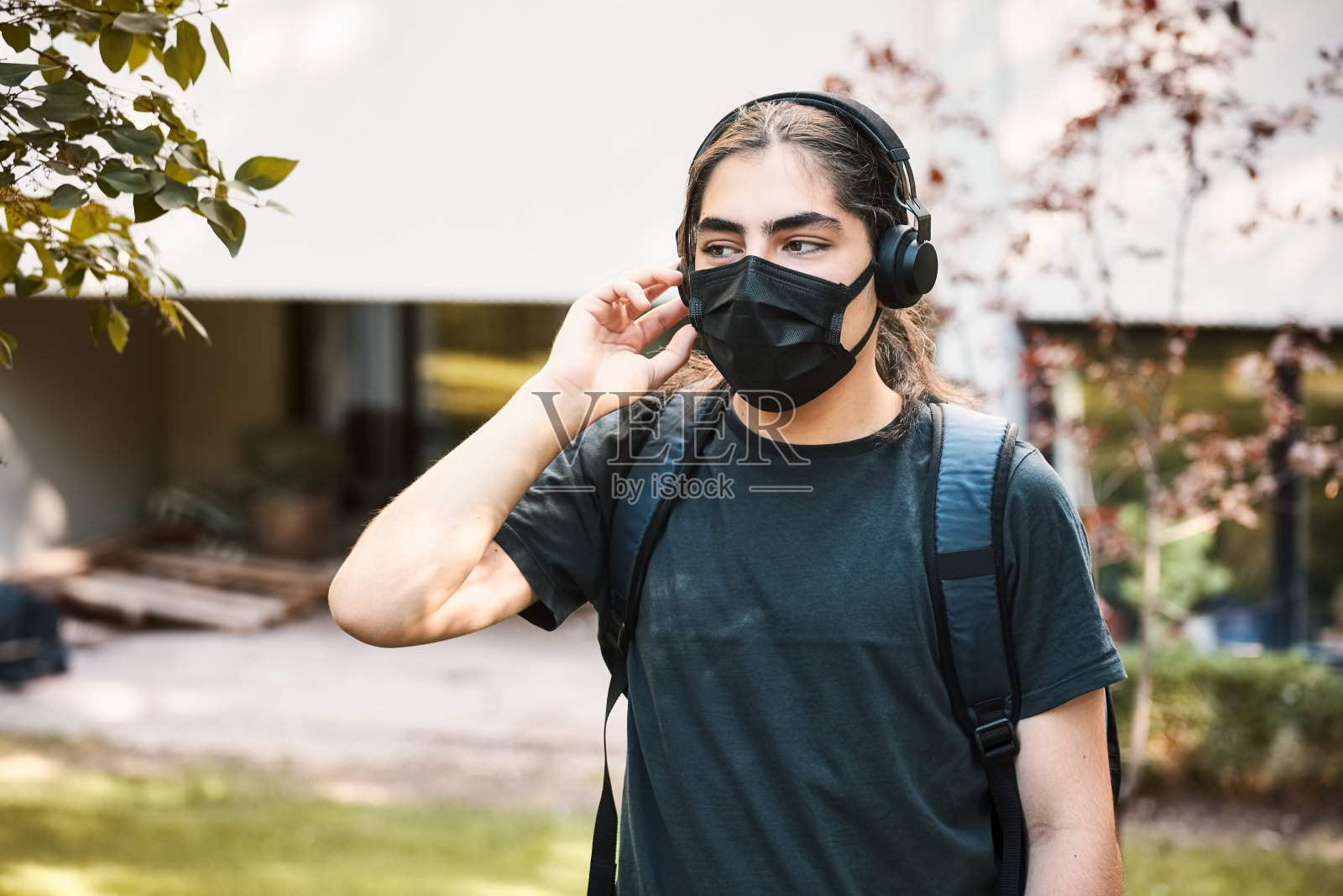 年轻的高中男生带着耳机和背包，戴着医用口罩，在学校的花园里听音乐。照片摄影图片