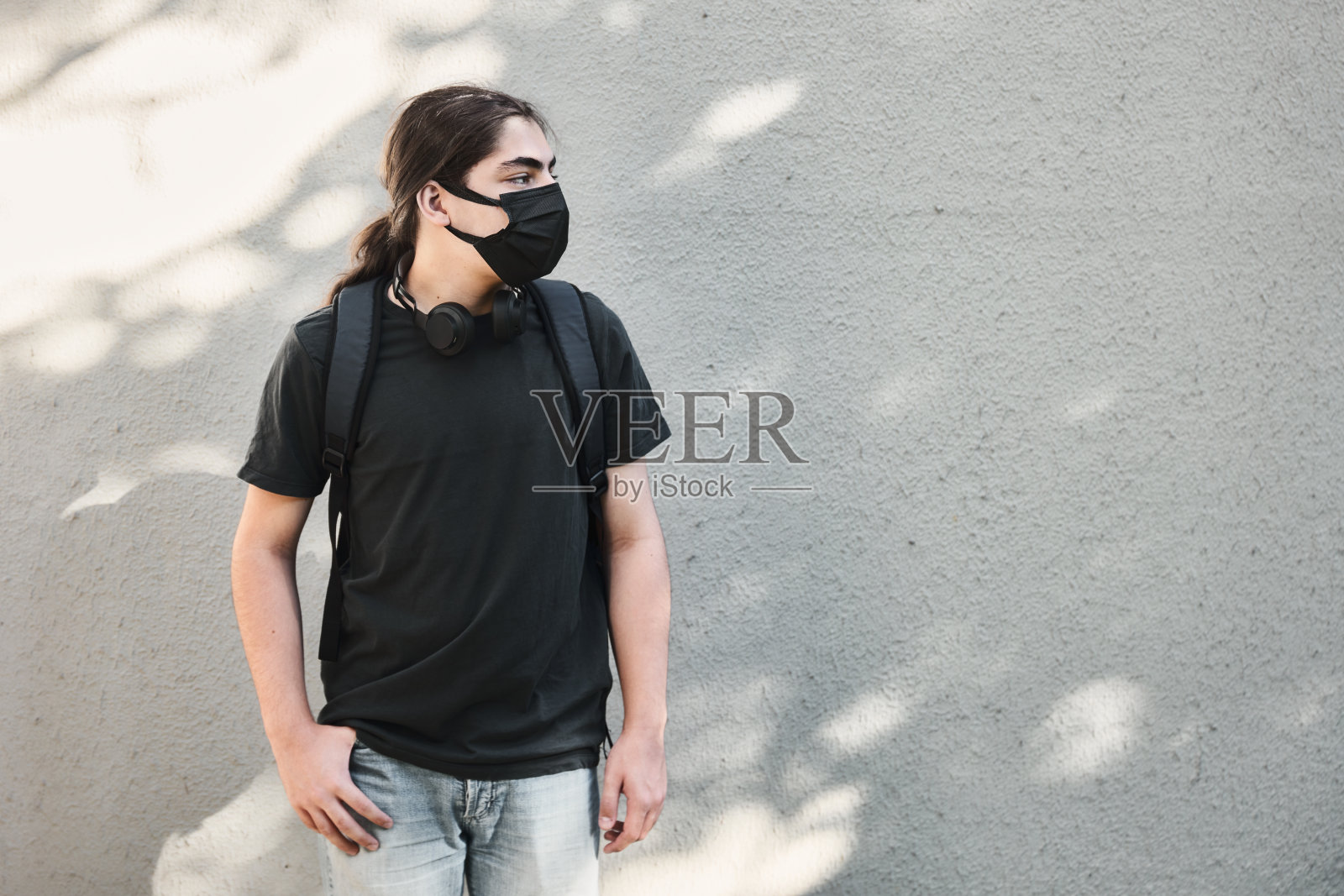 年轻的少年高中男孩带着耳机和背包，戴着白色墙背景的医用口罩照片摄影图片