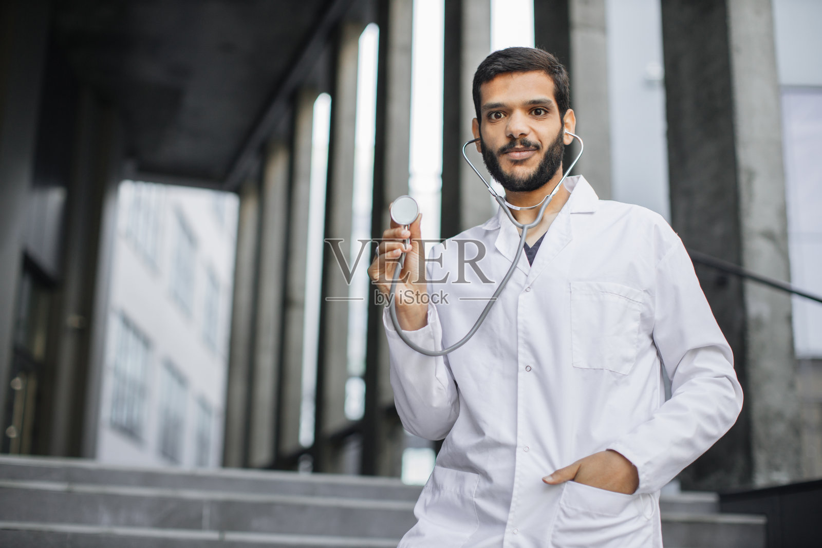 微笑的阿拉伯男医学生身穿白大褂，站在外面展示他的听诊器照片摄影图片