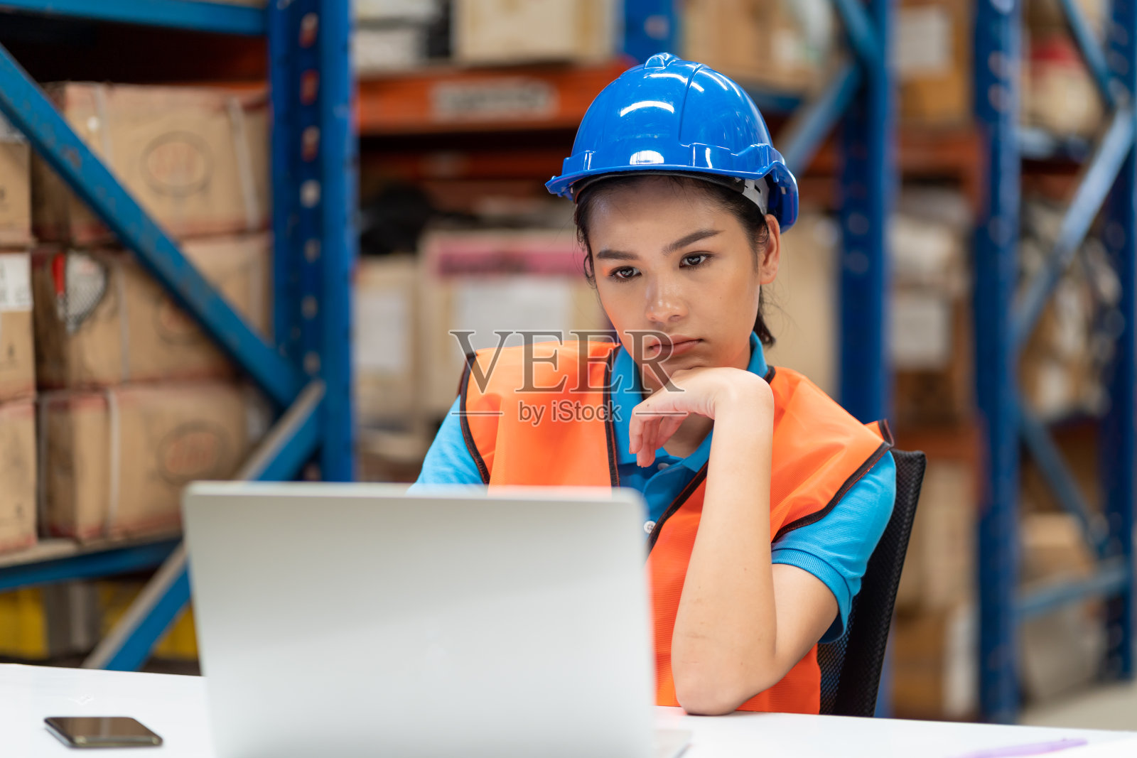 亚洲年轻女工人穿着安全背心和安全帽坐在工厂仓库的笔记本电脑上工作。人、仓库和行业概念照片摄影图片