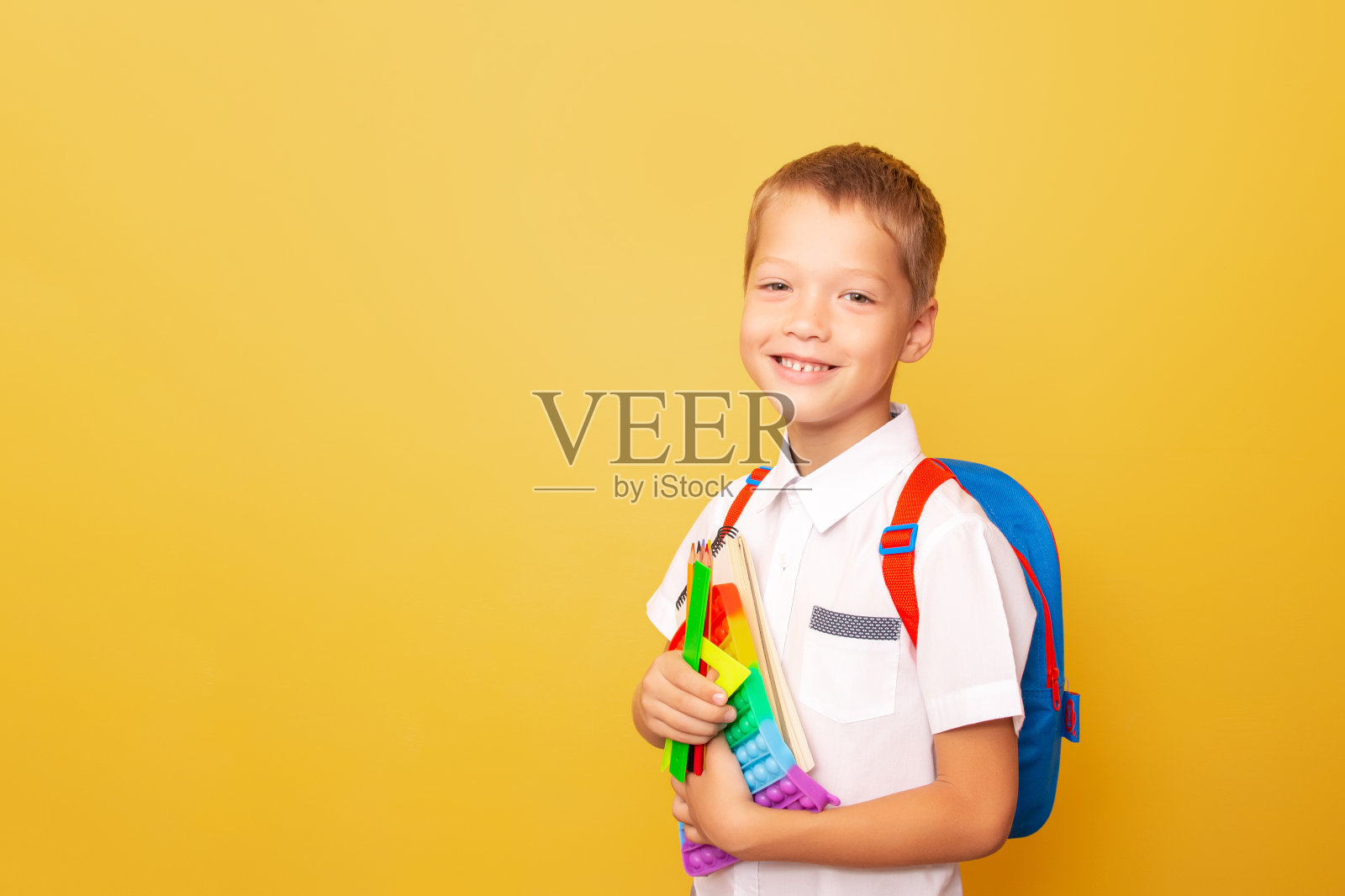 滑稽开朗微笑的白人男孩的肖像，白色衬衫与背包，手持文具和pop它，在黄色背景。学校教育理念，文具店。回到学校照片摄影图片