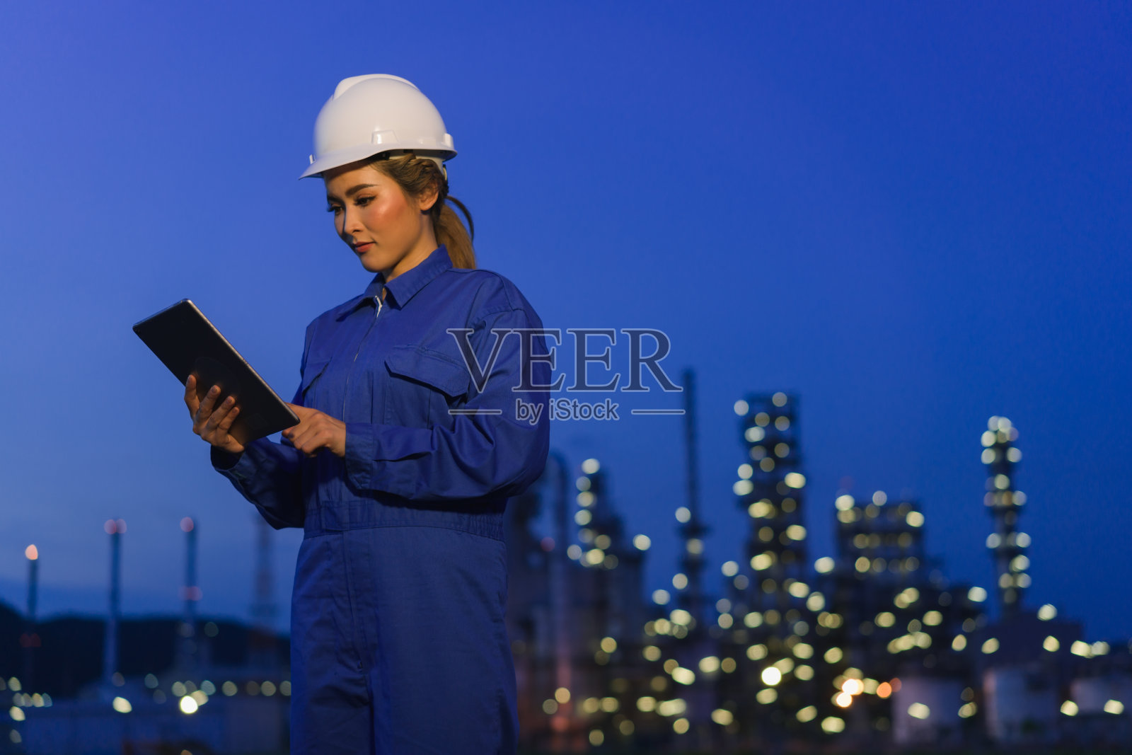 女工程师戴着白色安全帽，穿着女式套装，坐在石化工业门前看着平板电脑工作照片摄影图片