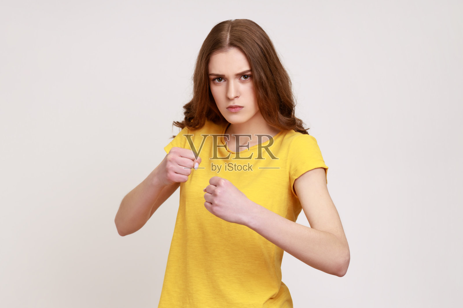 棕色头发的坚定少女穿着休闲的黄色t恤挥拳、拳击，自信地看着镜头。照片摄影图片