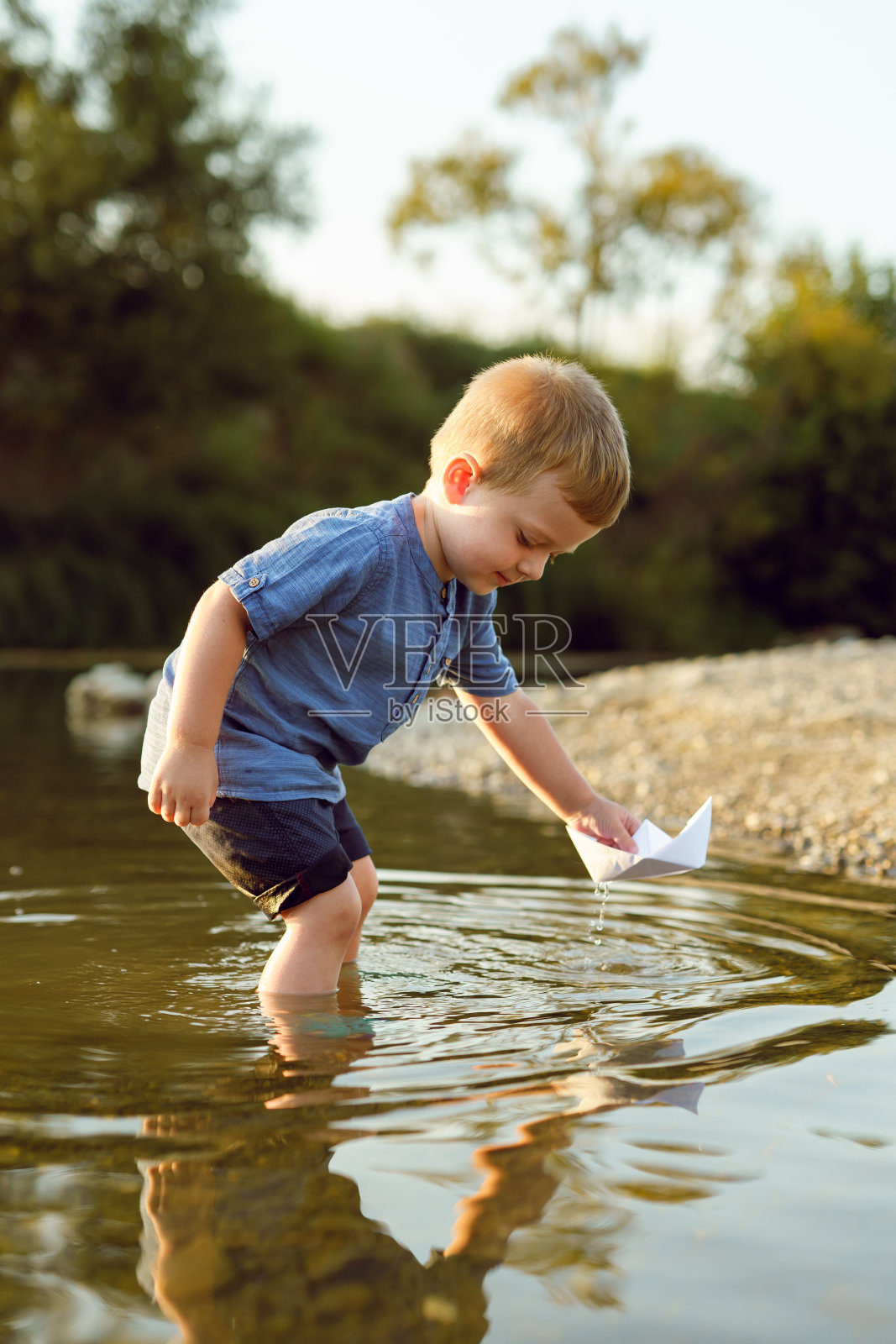 小白种人男孩，金发三岁，独自站在河边或湖边，在阳光明媚的夏天和秋天的傍晚玩着纸船童年宁静的概念复制空间照片摄影图片