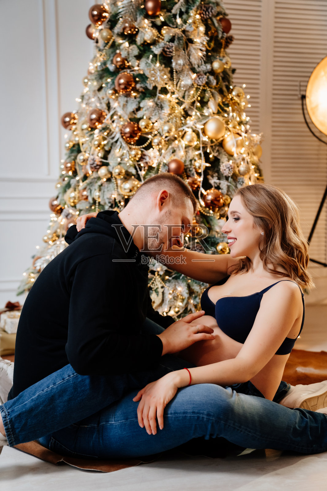 幸福的男人和怀孕的女人坐在圣诞树旁的地板上。照片摄影图片