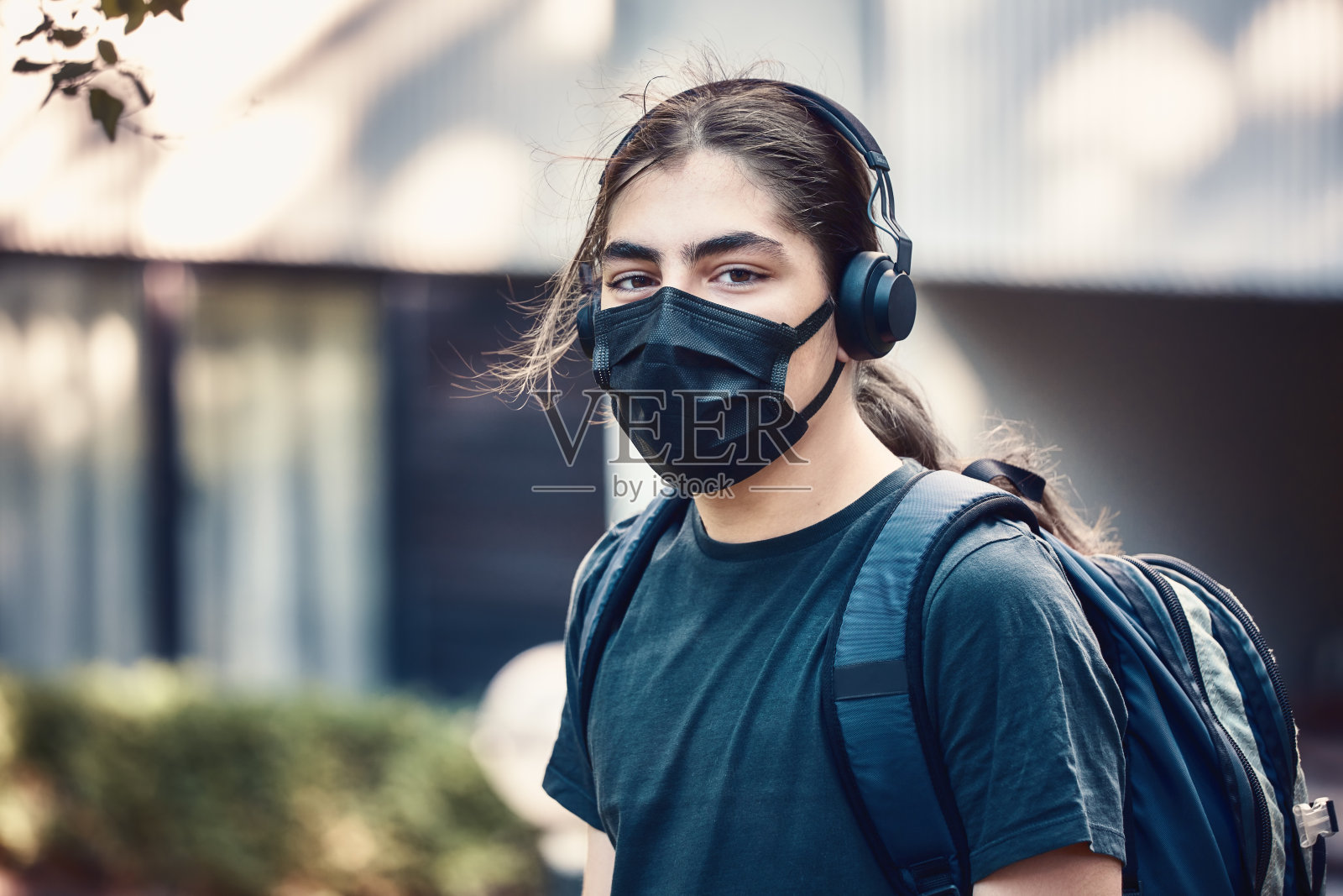 年轻的青少年或十几岁的高中男孩带着耳机和背包，戴着医用口罩在学校花园。照片摄影图片