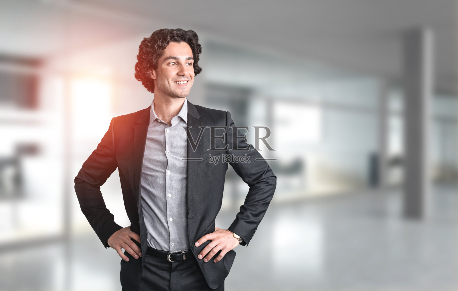 年轻英俊的商人穿着正式的黑色西装，双手叉腰站着，面带微笑，神情自信。模糊的全景办公室内部的背景。CEO和企业领导者的概念照片摄影图片