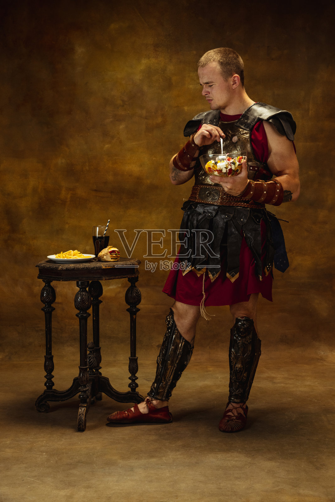 中世纪人的肖像，年轻人，战士或骑士在战争装备吃孤立在古老的黑暗背景。比较时代，艺术，历史，幽默，复古风格的概念照片摄影图片