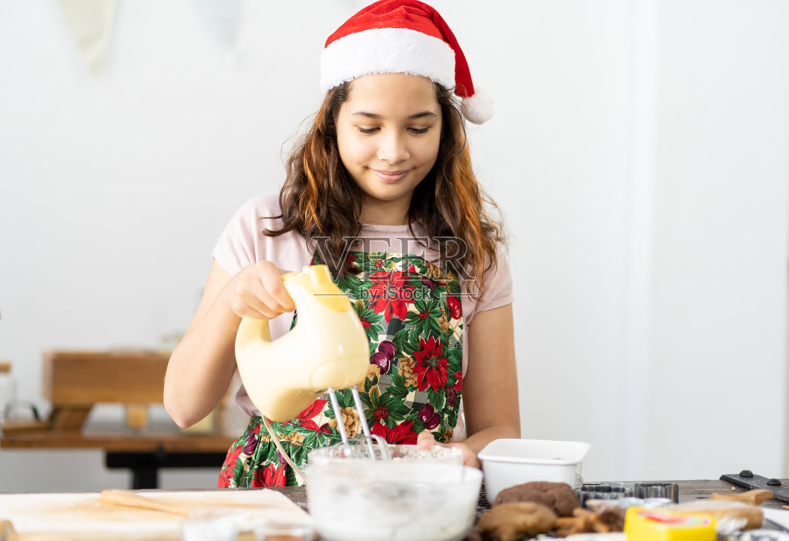 快乐的女孩戴着圣诞帽在家中厨房用手持搅拌机烹饪，圣诞快乐，新年快乐，烹饪和节日概念照片摄影图片