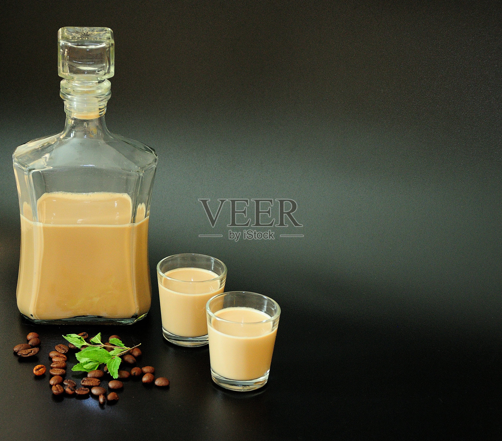 一瓶和两杯奶油咖啡利口酒，旁边是一把烤咖啡豆和一小枝新鲜薄荷，背景是黑色。照片摄影图片