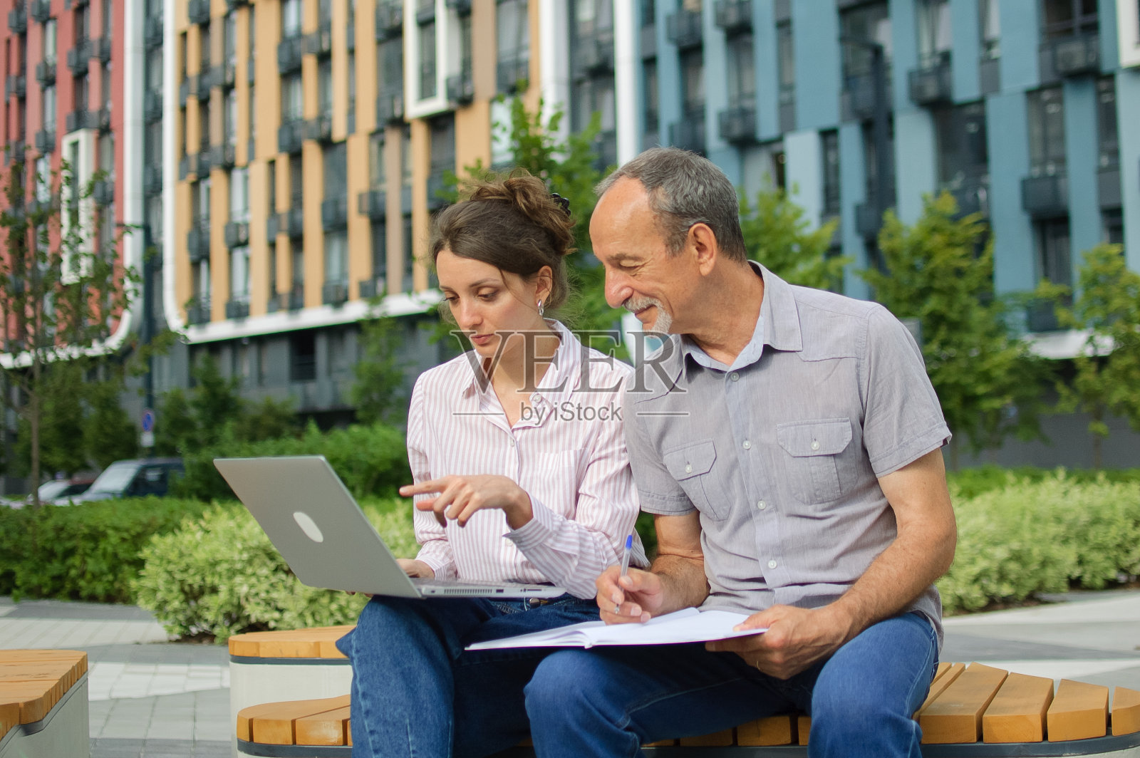 年轻貌美的女子和年长的男子正坐在现代住宅区的长椅上使用笔记本电脑。办公室外的非正式会议。一个女儿正在为她的父亲教授技术照片摄影图片
