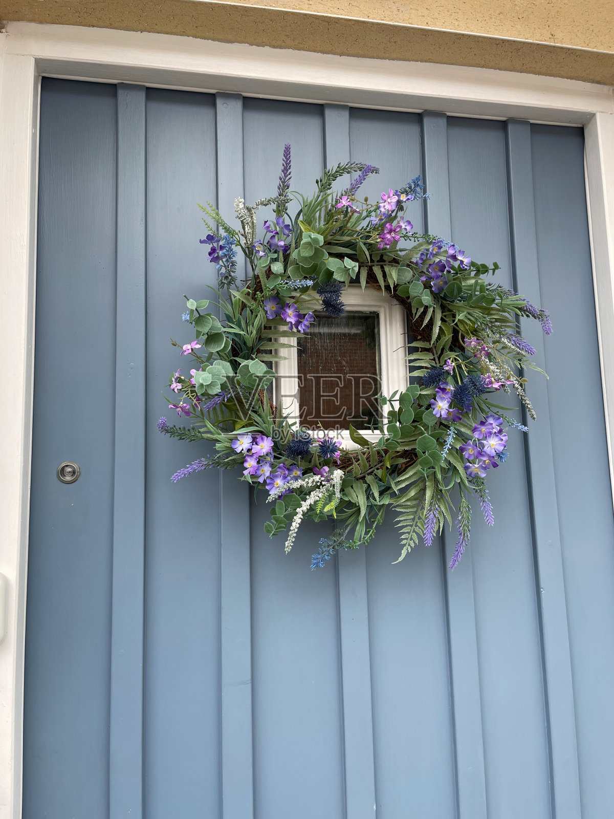 淡蓝的特写图像，木制镶板前门与眺望窗外和夏季花环装饰紫色，丝绸花和塑料，绿色树叶照片摄影图片