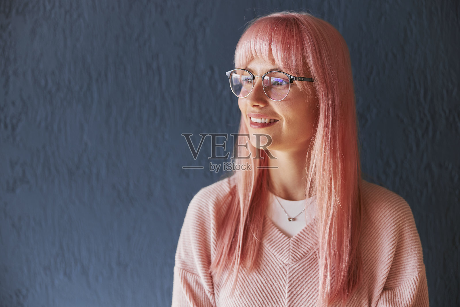 一个漂亮的女人，粉红色的头发，戴着眼镜，穿着毛衣站在蓝色的墙旁边照片摄影图片