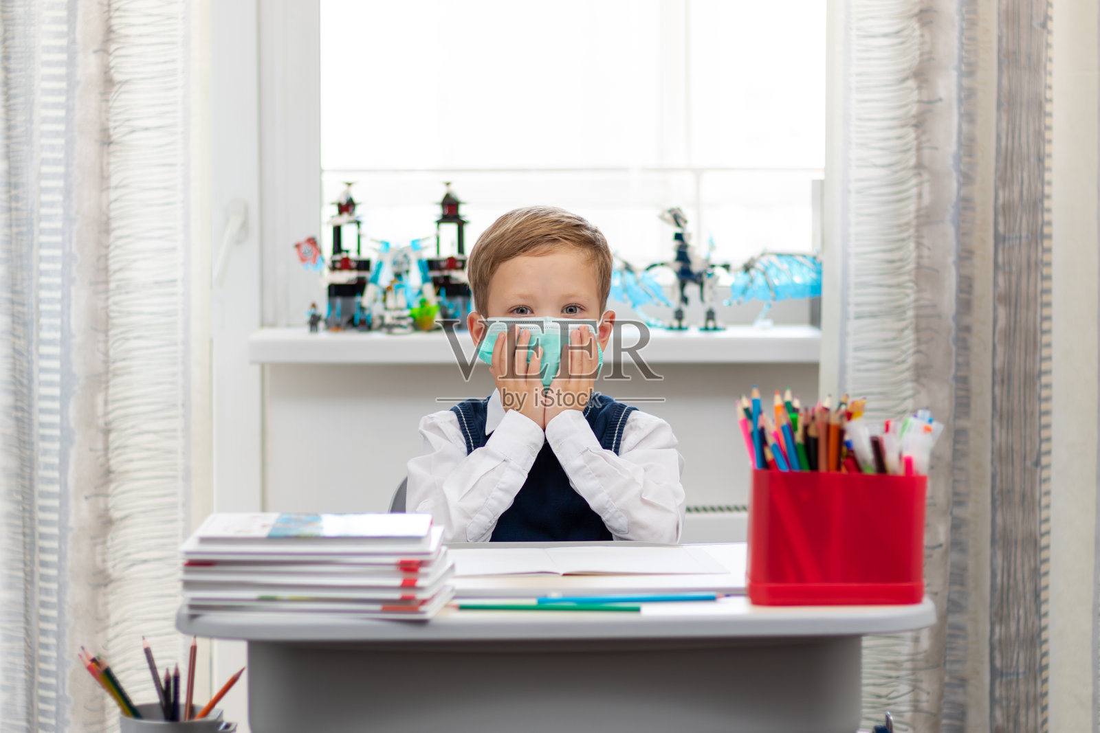 在冠状病毒大流行期间，一名可爱的一年级男孩穿着校服，戴着防护口罩坐在办公桌前。有选择性的重点。特写镜头照片摄影图片