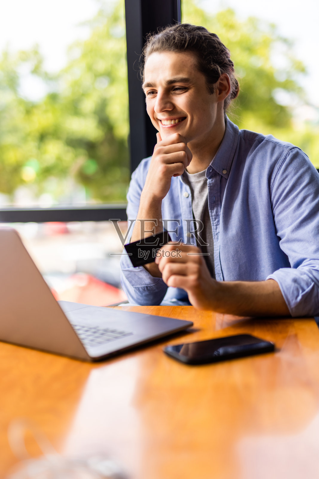 一个年轻人坐在咖啡馆里，手里拿着借记卡，手里拿着笔记本电脑。照片摄影图片