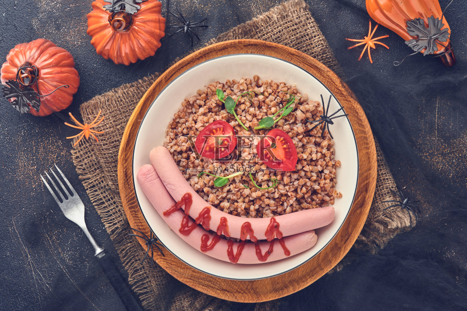 万圣节食品艺术创意儿童健康午餐。煮过的荞麦粥、肉香肠、西红柿和豌豆放在白色盘子里，供孩子们在石头背景下吃早餐或午餐。前视图。照片摄影图片