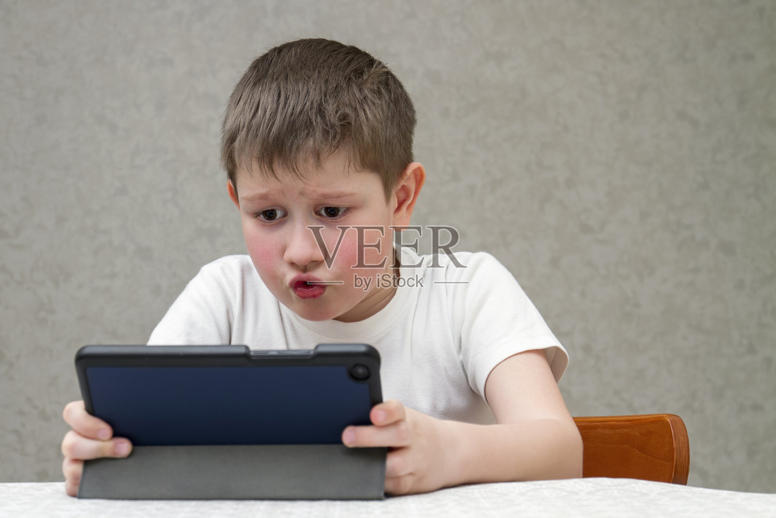 男孩情绪激动地玩着电子平板电脑。男孩带着平板坐在椅子上，放在家里的室内背景上。快乐的童年。积极的情绪和能量照片摄影图片