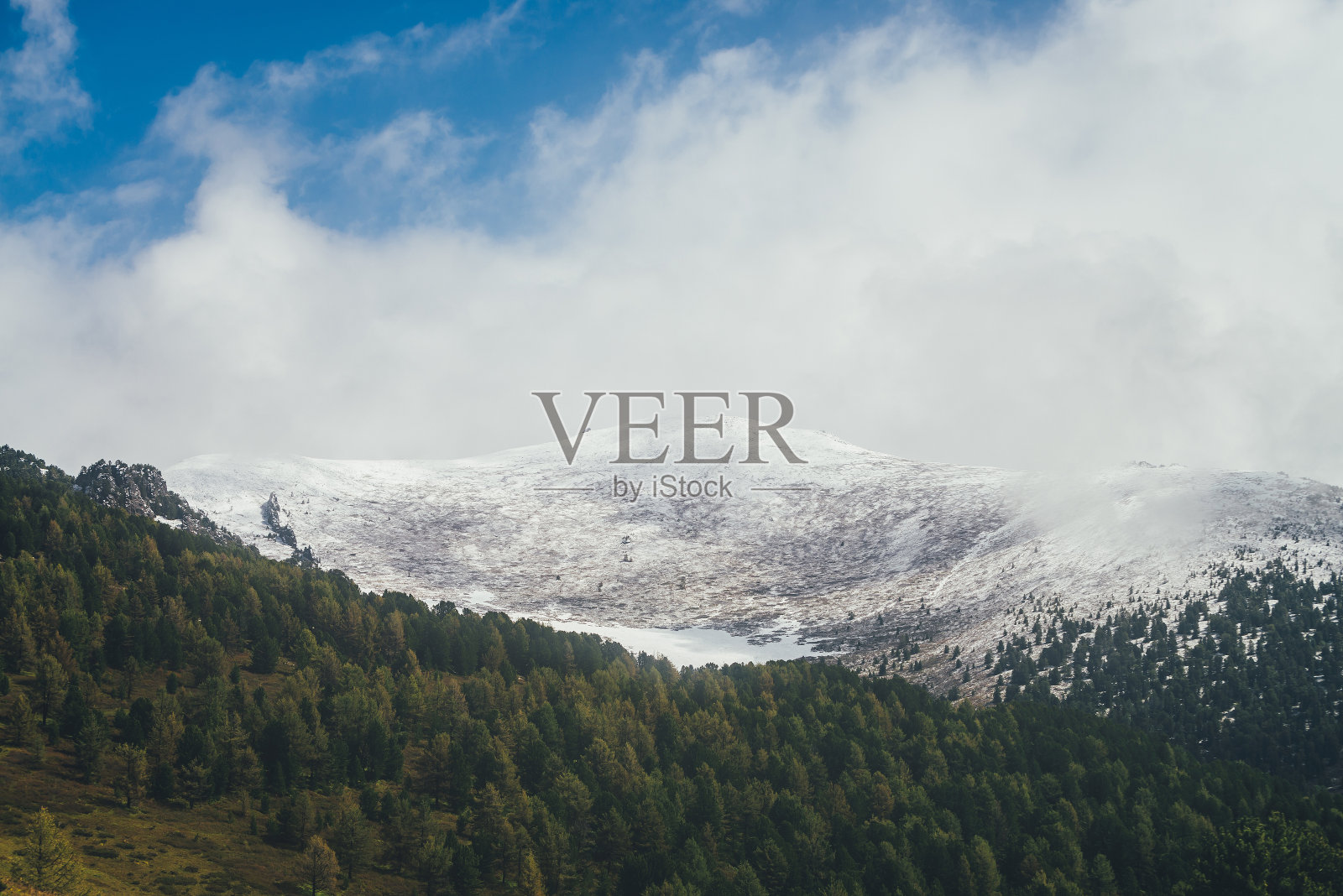 大气秋季景观与针叶林背景高积雪覆盖山顶低云层。令人敬畏的山景与绿色的森林和雪白的山峰在白云和蓝天照片摄影图片