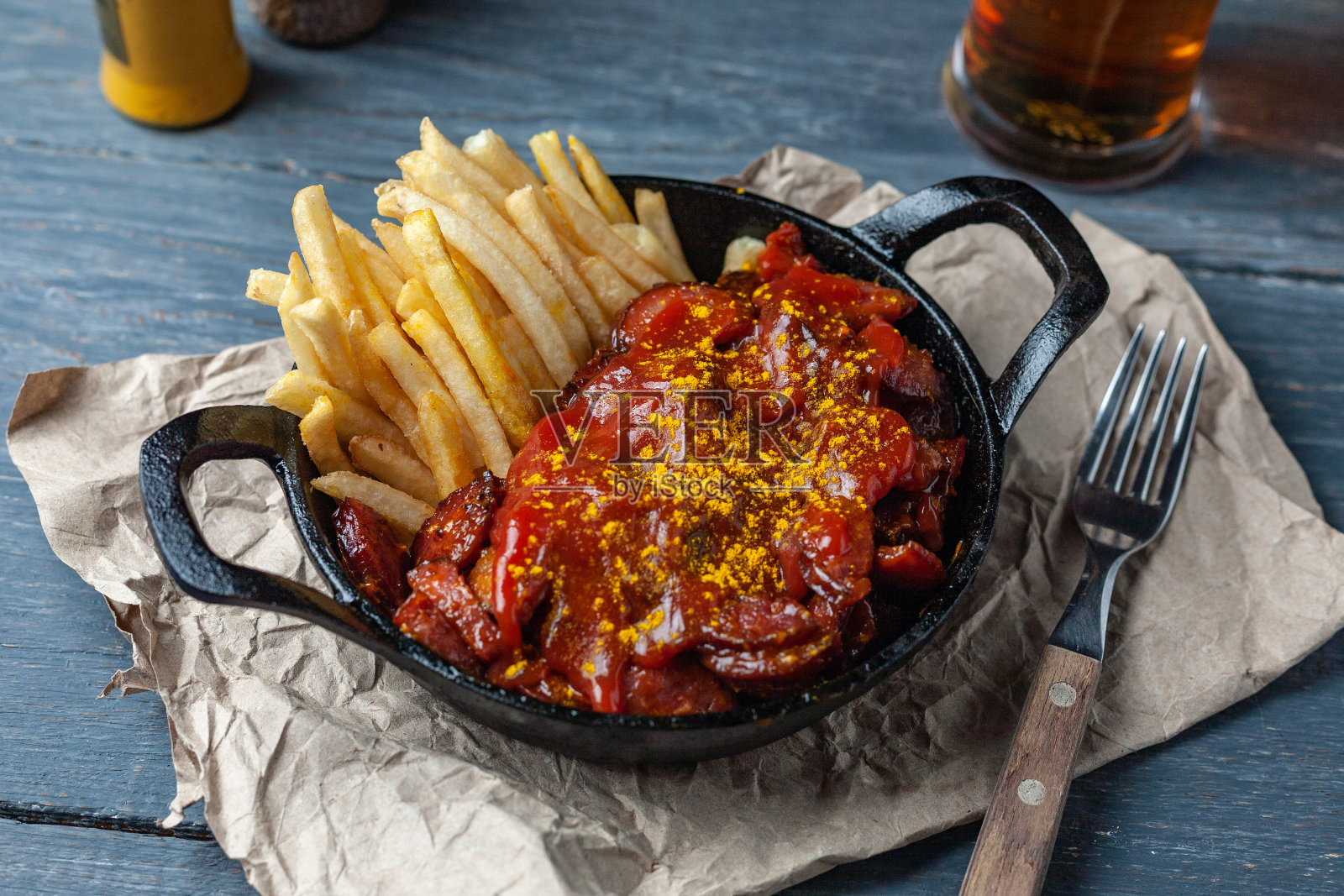 黑色平底锅里放着咖喱香肠和炸薯条，木桌上放着啤酒。照片摄影图片