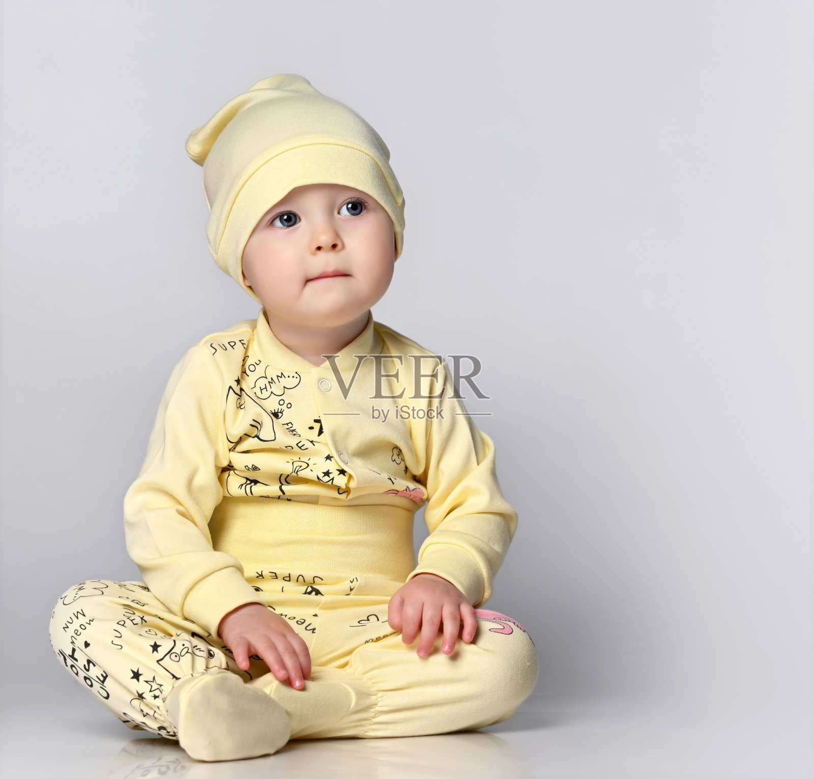 一个穿着长袖和shianishka的婴儿坐在工作室的地板上，他戴着一顶配套的帽子，衣服上有一个有趣的孩子气的图案照片摄影图片