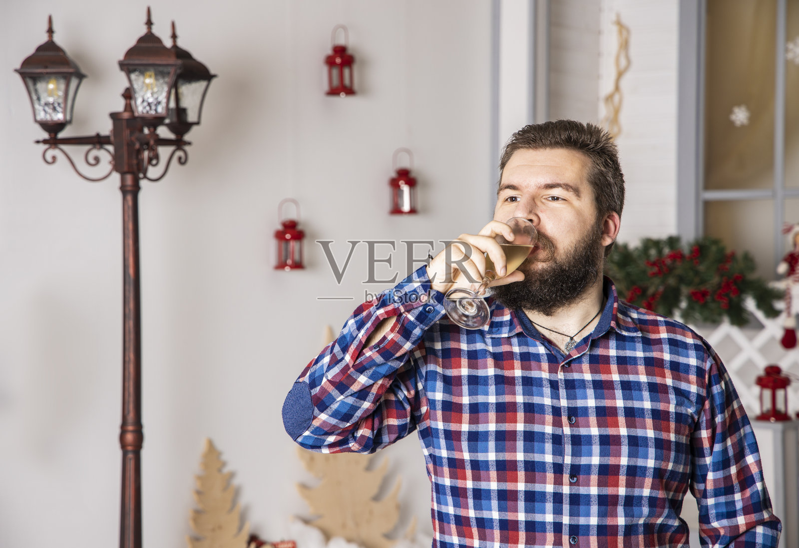 中年男子大胡子肖像在格子衬衫喝香槟从玻璃假日，白色室内工作室与街灯背景照片摄影图片